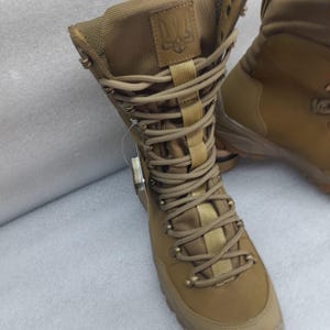 May include: Tan leather hiking boots with a brown lace-up closure and a black and yellow emblem on the side. The boots have a rugged sole and a high top.