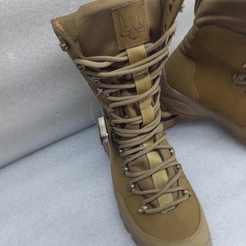 May include: Tan leather hiking boots with a brown lace-up closure and a black and yellow emblem on the side. The boots have a rugged sole and a high top.