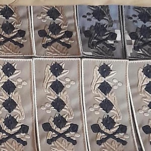 Puede incluir: Una colección de parches de tela beige con diseños bordados. Los parches presentan una imagen estilizada de un árbol con hojas y un símbolo de calavera y huesos cruzados. Los parches están dispuestos en filas y columnas.