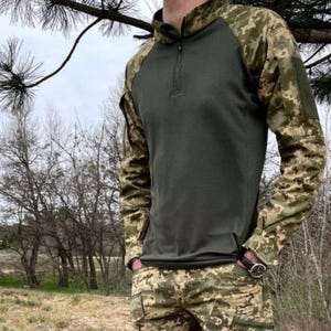 Può includere: Una camicia a maniche lunghe con un corpo verde scuro e maniche mimetiche. Le maniche hanno un motivo marrone, verde e beige.