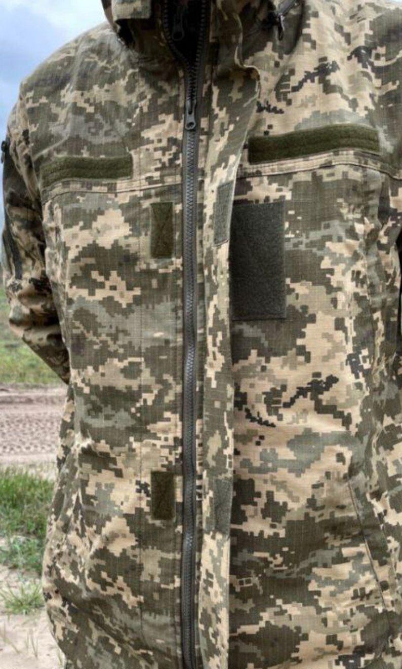 May include: A camouflage jacket with a full-length zipper and a hood. The jacket features a digital camouflage pattern in shades of green, brown, and tan. It has multiple pockets and patches, and is designed for outdoor use.