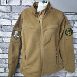 May include: A brown fleece jacket with a zip fastening and two pockets. The jacket has a patch on the left sleeve with a skull and crossbones and the word "BOZIH" written below it. There is another patch on the right sleeve with a woman in a bikini and the words "THE  REVENGE" written below it.