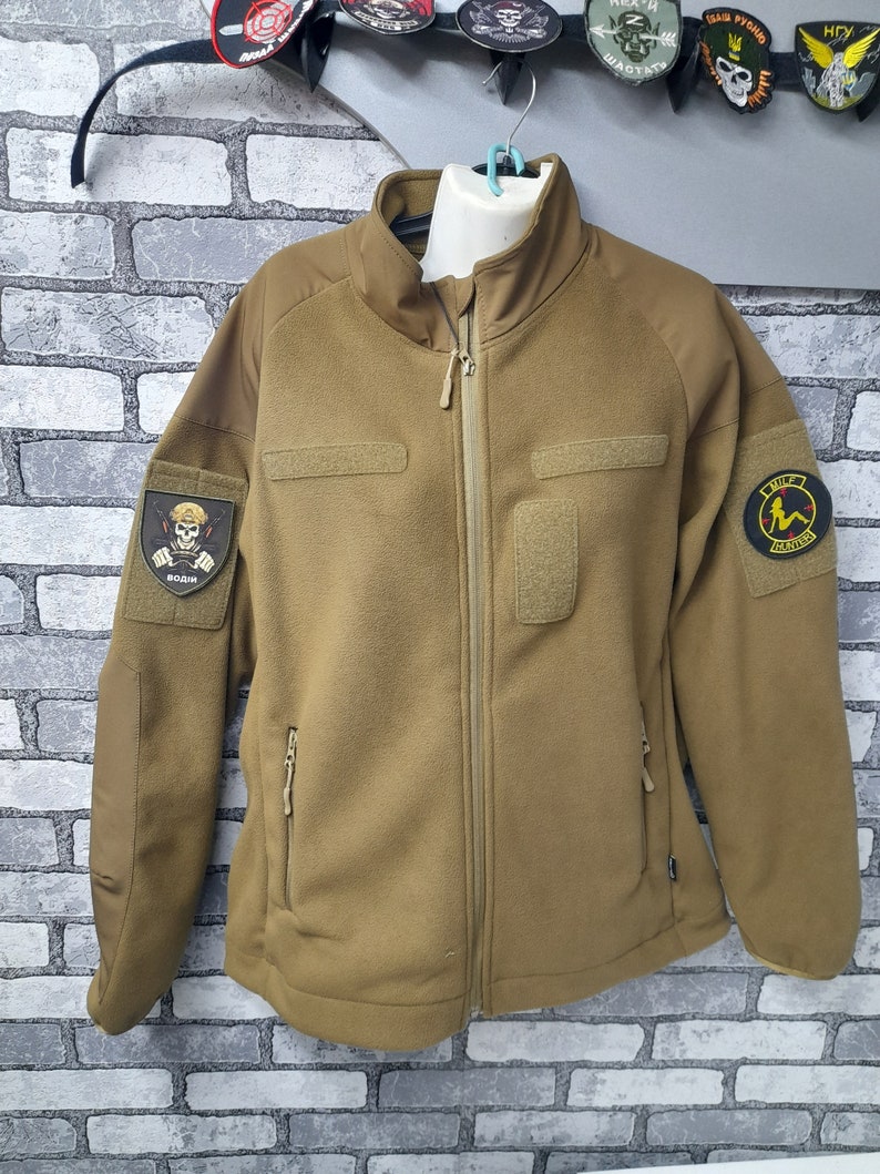 May include: A brown fleece jacket with a zip fastening and two pockets. The jacket has a patch on the left sleeve with a skull and crossbones and the word "BOZIH" written below it. There is another patch on the right sleeve with a woman in a bikini and the words "THE  REVENGE" written below it.