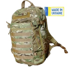 Könnte beinhalten: Ein Tarnrucksack mit mehreren Fächern und verstellbaren Riemen. Der Rucksack hat einen oberen Griff und verstellbare Schultergurte. Der Text "PTNPNX" ist auf einem Aufnäher zu sehen. Ein Etikett lautet "MADE IN UKRAINE".