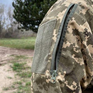 May include: Close-up of a camouflage jacket sleeve. The jacket features a digital pixelated pattern in shades of green, brown, and tan. A gray patch is sewn onto the upper arm, and a long, dark zipper runs down the sleeve.