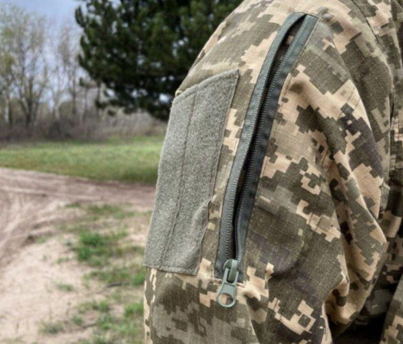May include: Close-up of a camouflage jacket sleeve. The jacket features a digital pixelated pattern in shades of green, brown, and tan. A gray patch is sewn onto the upper arm, and a long, dark zipper runs down the sleeve.