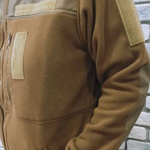 Puede incluir: Una chaqueta de forro polar marr&oacute;n con cierre de cremallera y dos bolsillos. La chaqueta tiene un parche marr&oacute;n en el hombro izquierdo y un parche marr&oacute;n en la manga derecha.