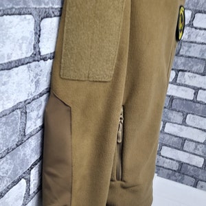 May include: A brown fleece jacket with a zipped pocket on the left side. The jacket has a patch on the left shoulder and a small patch on the right side.