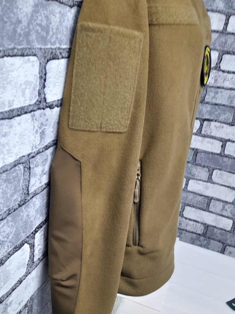 May include: A brown fleece jacket with a zipped pocket on the left side. The jacket has a patch on the left shoulder and a small patch on the right side.