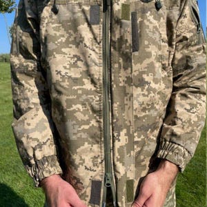 May include: A camouflage jacket in shades of tan, green, and brown. The jacket features a full-length zipper, multiple pockets, and a hood. The sleeves have elastic cuffs. The jacket is designed for outdoor wear.