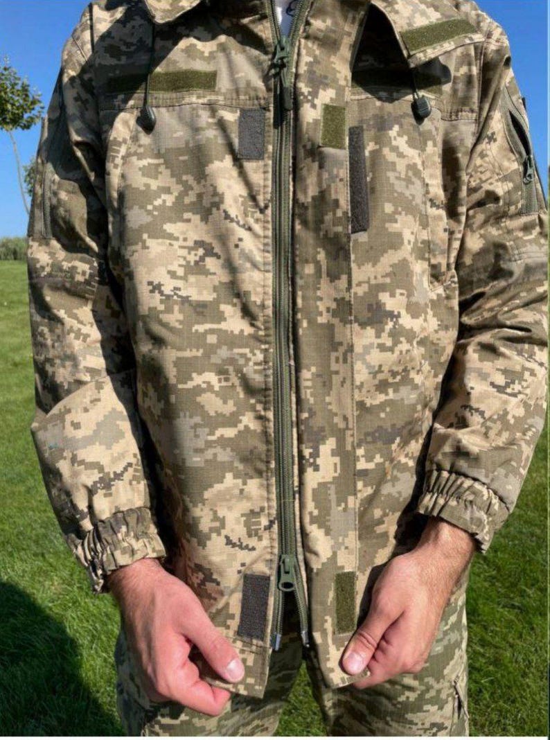 May include: A camouflage jacket in shades of tan, green, and brown. The jacket features a full-length zipper, multiple pockets, and a hood. The sleeves have elastic cuffs. The jacket is designed for outdoor wear.