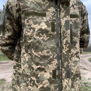 May include: A camouflage jacket in shades of green, brown, and tan. The jacket features a full-length zipper, multiple pockets, and patches. The fabric has a pixelated pattern, typical of military-style clothing.