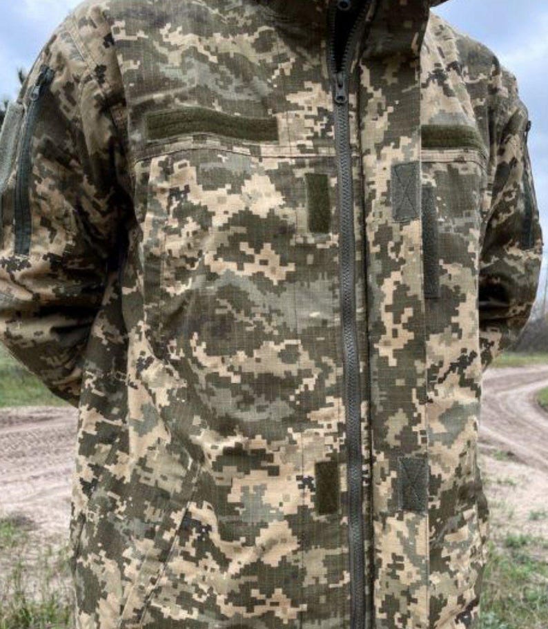 May include: A camouflage jacket in shades of green, brown, and tan. The jacket features a full-length zipper, multiple pockets, and patches. The fabric has a pixelated pattern, typical of military-style clothing.