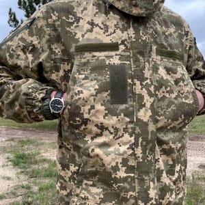 Könnte beinhalten: Eine Tarnjacke in Grün-, Braun- und Beigetönen. Die Jacke hat eine Kapuze, mehrere Taschen und eine schwarze Uhr. Das pixelartige Muster ist ein wichtiges Designelement.