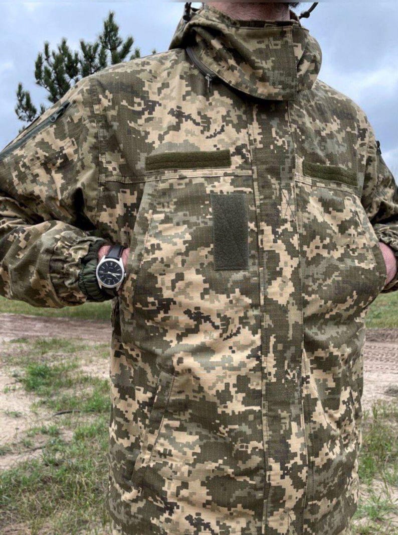 A camouflage jacket in shades of green, brown, and tan. The jacket has a hood, multiple pockets, and a black watch on the wrist. The jacket is designed with a pixelated pattern.