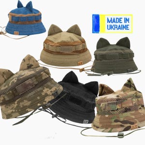 May include: A collection of cat ear bucket hats in various colors and patterns. The hats include denim blue, tan, olive green, camouflage, and black. Each hat features a mesh panel and adjustable chin strap. The hats are labeled "Made in Ukraine".