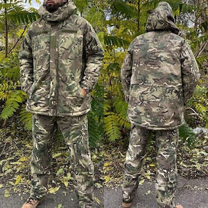 May include: A camouflage jacket and trousers set in green, brown, and beige. The jacket features a hood and pockets, and the trousers have pockets on the legs. The outfit is designed for outdoor activities and blends with natural surroundings.