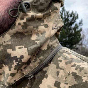 May include: Close-up of a camouflage jacket with a hood. The jacket features a digital camouflage pattern in shades of green, tan, and brown. A zipper runs diagonally across the chest. The hood has a drawstring and a toggle for adjustment.