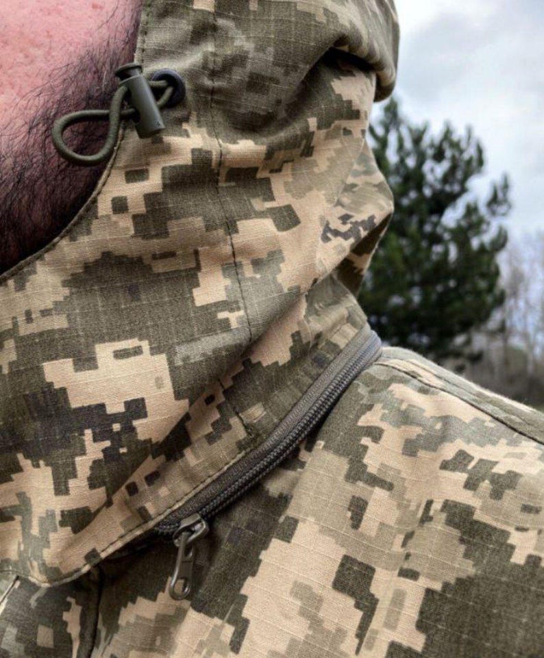 May include: Close-up of a camouflage jacket with a hood. The jacket features a digital camouflage pattern in shades of green, tan, and brown. A zipper runs diagonally across the chest. The hood has a drawstring and a toggle for adjustment.