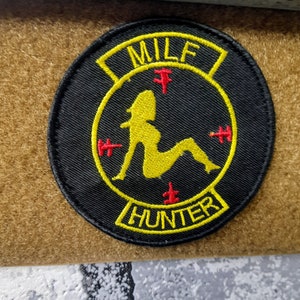 Op de afbeelding: Een geborduurde zwarte en gele patch met de tekst "MILF HUNTER" en een silhouet van een vrouw in zittende positie.