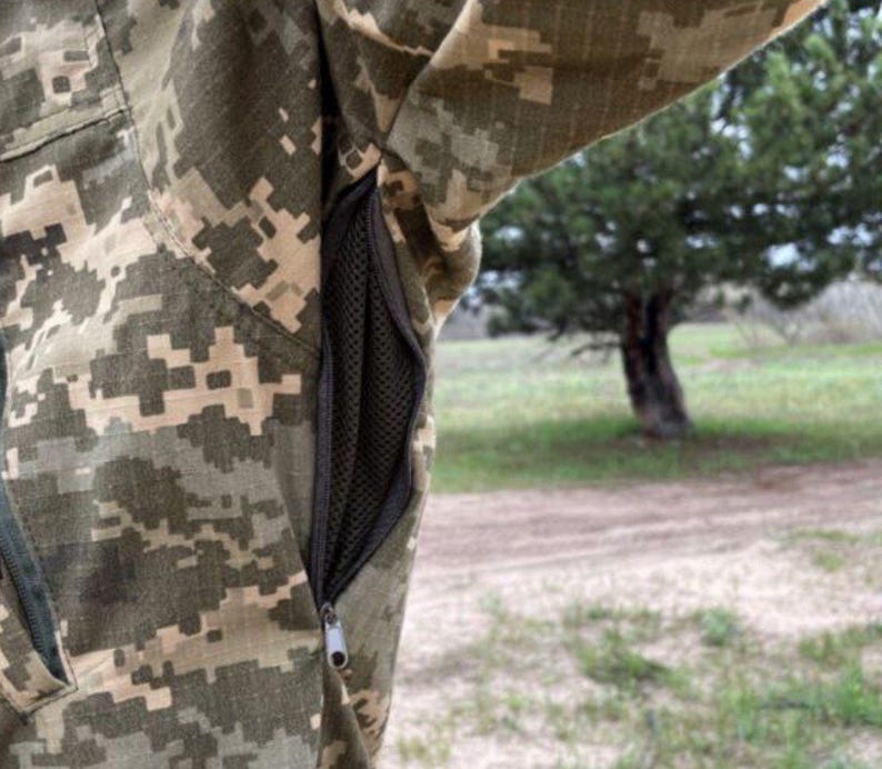May include: Close-up of a camouflage jacket with a digital pixel pattern in shades of green, tan, and brown. The jacket features a black mesh pocket with a zipper closure. The background shows a blurred outdoor scene with a tree.