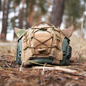 May include: An olive green tactical helmet with a camouflage cover and tan detailing. The helmet has a mesh front and various straps and attachments. The design includes two horn-like protrusions on the sides, suggesting a military or tactical theme.