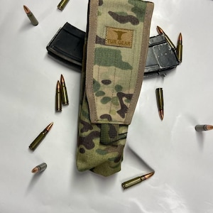 Peut inclure: Un étui de chargeur à motif camouflage avec le texte "TUR GEAR" et plusieurs balles éparpillées autour. L'étui est de couleur beige, marron et vert. Deux chargeurs noirs sont également visibles.