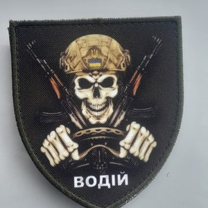 May include: A green, shield-shaped patch with a skull wearing a military helmet and driving a steering wheel. The skull is holding two rifles crossed behind it. The text "ВОДИЙ" is printed below the skull.