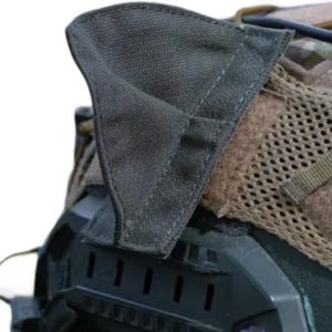 Puede incluir: Primer plano de un casco táctico con exterior verde oscuro y patrón de camuflaje. El casco presenta una solapa de tela verde oscuro y una sección de malla. La parte inferior del casco es negra, con una serie de pequeñas aberturas rectangulares.