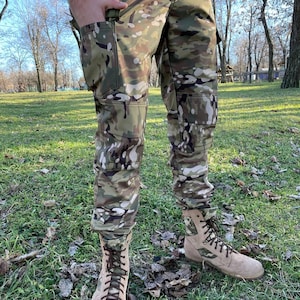 May include: Camouflage pattern pants and matching jacket with multiple pockets. The pants are paired with tan combat boots with dark brown laces. The outfit is designed for outdoor activities and features a military-inspired aesthetic.