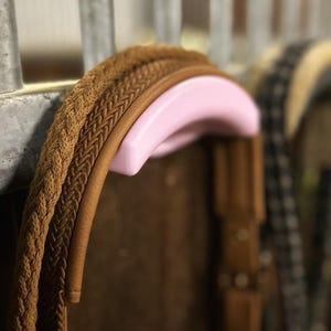 Könnte beinhalten: Nahaufnahme von Reitausrüstung, mit einem braunen geflochtenen Seil und einem Lederriemen, die an einem rosa gebogenen Halter hängen. Der Hintergrund ist unscharf, was auf eine Stallumgebung hindeutet. Der Fokus liegt auf den Details des Zaumzeugs.
