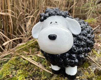Könnte beinhalten: Eine schwarze und weiße Keramik-Schaffigur mit einem Comic-Gesicht und schwarzem lockigem Wollfell. Das Schaf steht auf einem Bett aus Moos und trockenem Gras.