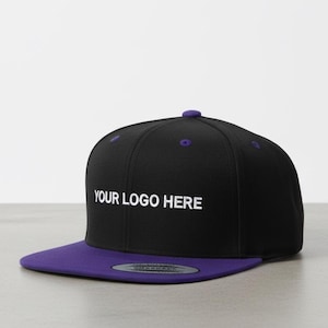 May include: A black snapback cap featuring a flat purple brim and a purple button on the crown. The front panel displays the white text YOUR LOGO HERE in a sans-serif font. A circular sticker is visible on the top surface of the flat brim.