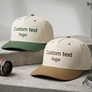 May include: Two baseball caps with the text "Custom text logo". One cap has a cream-colored crown and a green brim. The other cap has a cream-colored crown and a brown brim. The caps are on a concrete surface.