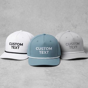 Può includere: Tre cappellini da baseball bianchi, verde acqua e grigi, ciascuno con "CUSTOM TEXT" stampato sul davanti. Il cappellino bianco ha un dettaglio in corda nera, mentre i cappellini verde acqua e grigi hanno dettagli in corda bianca.
