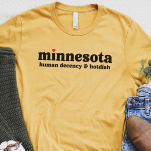 May include: A yellow t-shirt with the text "minnesota human decency & hotdish" printed on it. The shirt is laid out on a white surface with a pair of brown boots, blue jeans, and a green sweater.