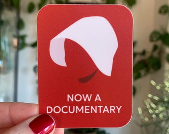 May include: Red sticker with a white silhouette of a woman wearing a white bonnet. The text "NOW A DOCUMENTARY" is printed in white below the silhouette.