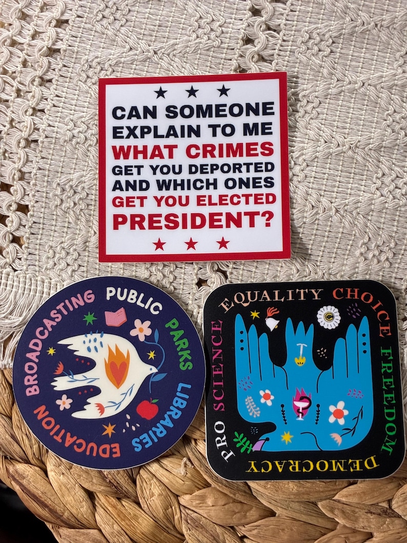 Op de afbeelding: Drie stickers met politieke en sociale rechtvaardigheidsthema's. E&eacute;n leest: "Kan iemand mij uitleggen welke misdaden ervoor zorgen dat je wordt gedeporteerd en welke ervoor zorgen dat je tot president wordt gekozen?" Een andere zegt "Broadcasting Public Parks Libraries Education". De derde zegt "Equality Choice Freedom Pro Science Democracy".