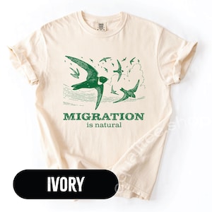 May include: Ivory-colored t-shirt with a green illustration of birds in flight and the text "MIGRATION is natural." The design features several birds soaring across the sky, with a detailed depiction of one prominent bird. The shirt's color is ivory.
