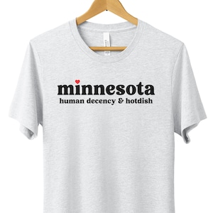 May include: A white t-shirt with black text that reads "minnesota human decency & hotdish" with a red heart above the word "minnesota".