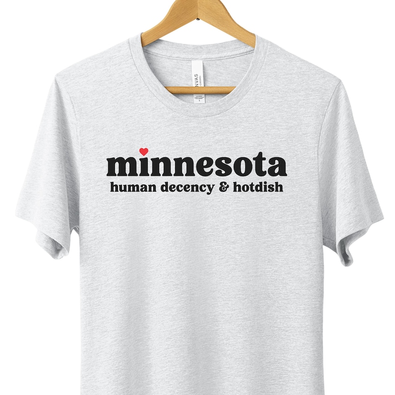 May include: A white t-shirt with black text that reads "minnesota human decency & hotdish" with a red heart above the word "minnesota".
