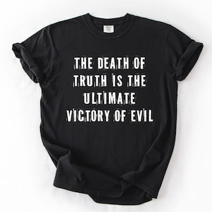 May include: Black t-shirt with white text. The text reads: "THE DEATH OF TRUTH IS THE ULTIMATE VICTORY OF EVIL." The shirt has a crew neck and short sleeves. The text is in a distressed, slightly blurred font.
