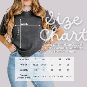 May include: A size chart for a Comfort Colors 3023CL heavyweight boxy t-shirt. The chart shows measurements in inches for width, length, and sleeve center back for sizes S, M, L, XL, and XXL. The t-shirt is a dark gray color.