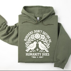 Pode incluir: Moletom verde-oliva com a frase branca "Borders Don't Define Us Humanity Does". O design inclui dois p&aacute;ssaros e elementos florais. O moletom tem capuz com cord&atilde;o e mangas compridas.