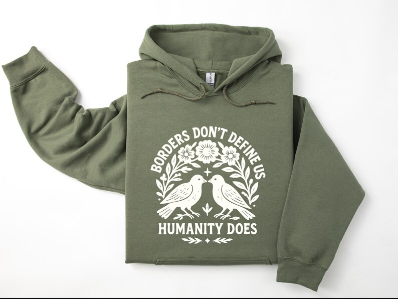 Pode incluir: Moletom verde-oliva com a frase branca "Borders Don't Define Us Humanity Does". O design inclui dois p&aacute;ssaros e elementos florais. O moletom tem capuz com cord&atilde;o e mangas compridas.