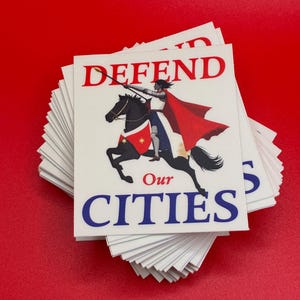 May include: A stack of white stickers with a graphic of a person on a black horse, holding a spear. The stickers have the words "DEFEND Our CITIES" in red and blue text.