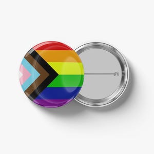 May include: A round, glossy button with a rainbow design and the Progress Pride flag. The button is red, orange, yellow, green, blue, and purple. The Progress Pride flag includes pink, light blue, brown, and black. A second button is visible.