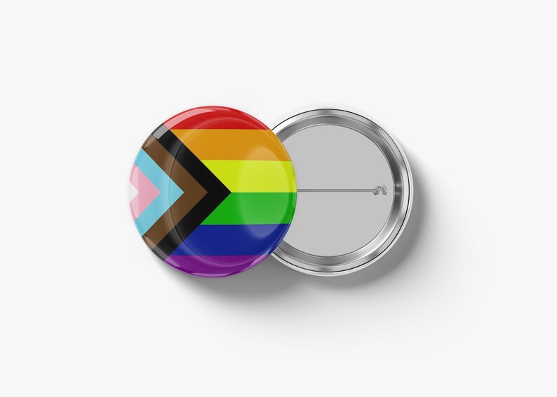 May include: A round, glossy button with a rainbow design and the Progress Pride flag. The button is red, orange, yellow, green, blue, and purple. The Progress Pride flag includes pink, light blue, brown, and black. A second button is visible.