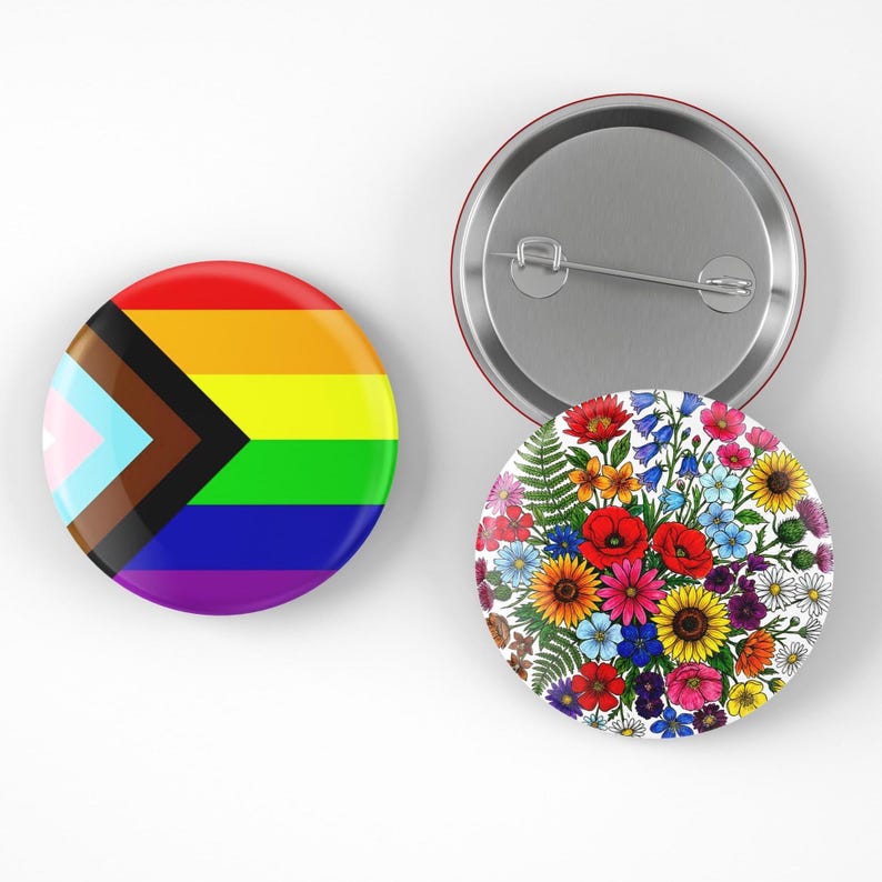 May include: Two round pinback buttons. One features the Progress Pride flag with rainbow stripes and a chevron of colors. The other has a floral design with red poppies, sunflowers, and other colorful flowers. The back of one button is visible.