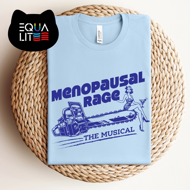 May include: Light blue t-shirt with a blue graphic of a woman sitting on a chainsaw with the text "Menopausal Rage - The Musical".